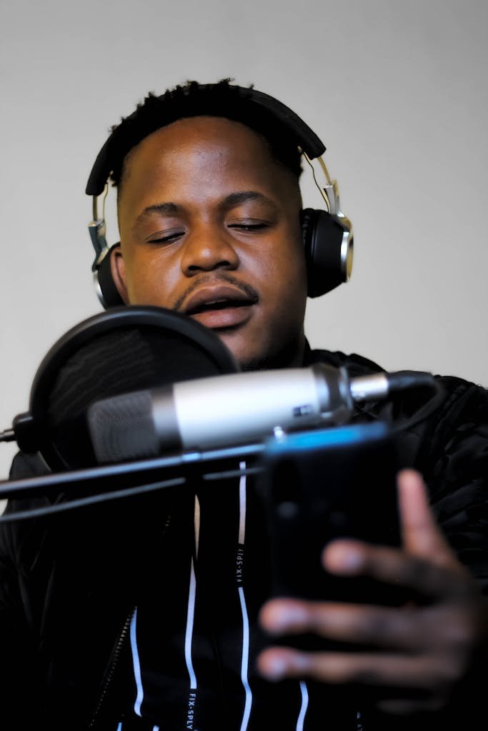 A close-up Lyrics portrait of a man singing into a microphone, wearing headphones, and holding a cellphone.