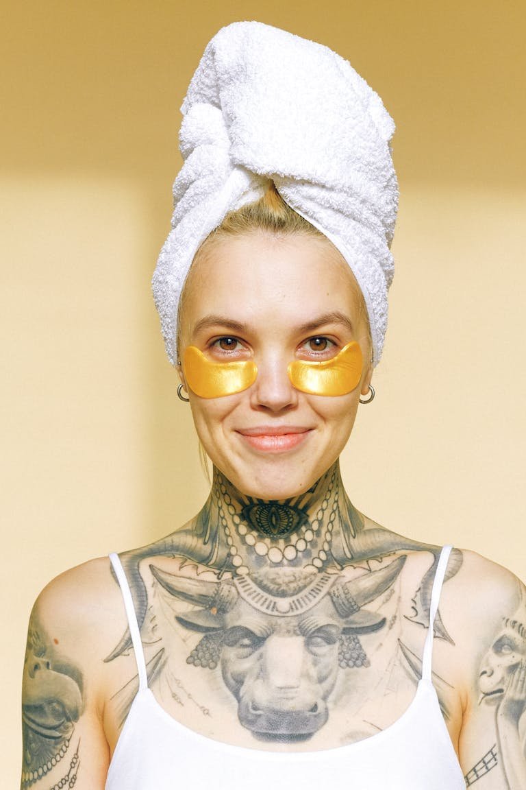 A woman Beauty Tips – Well Health Organic.com with tattoos, towel on head, and skincare patches smiles warmly.