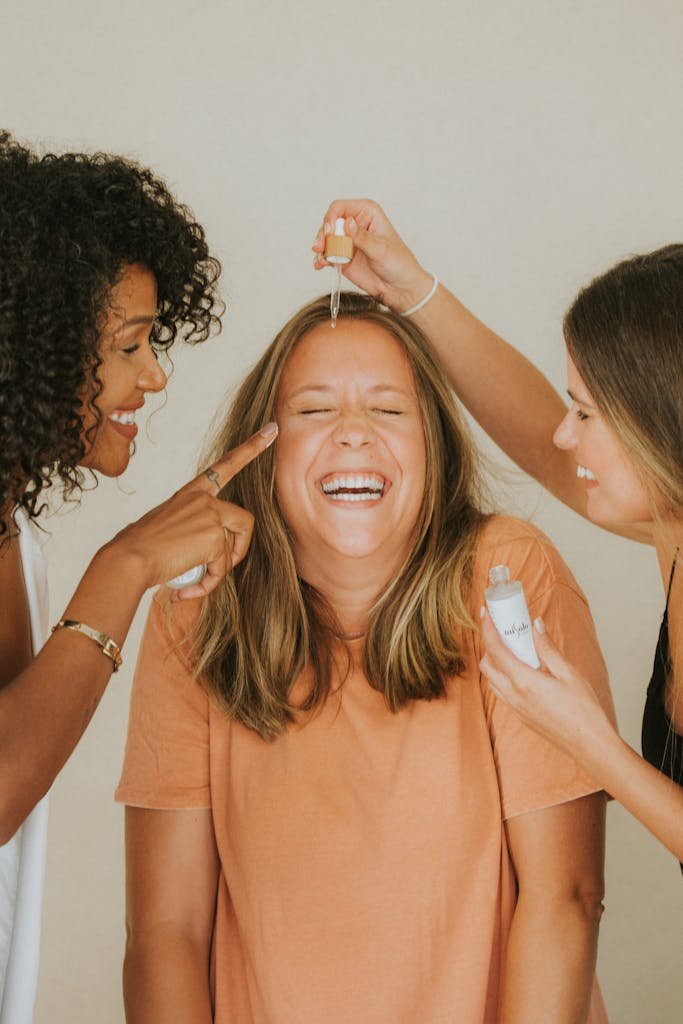 Three womenBeauty Tips – Well Health Organic.com laughing and applying skincare products indoors.