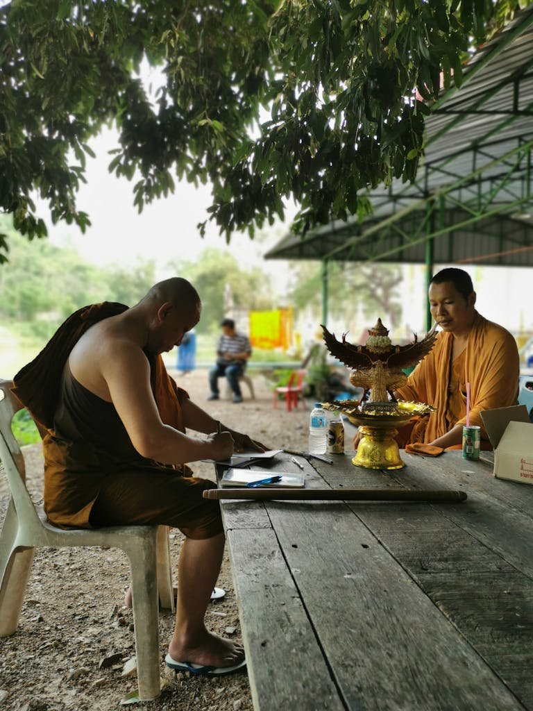 Linawang26 Two monks sitting at a table under a tree in Tambon Wang Lum, Thailand, embracing tranquility.