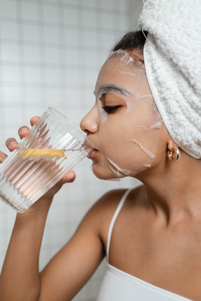 Woman in skincare Beauty Tips – Well Health Organic.com routine with sheet mask, drinking lemon water for hydration.